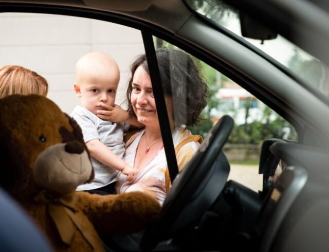 donna con un bambino in braccio che guardano dentro ad una macchina con un orsacchiotto alla guida
